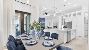 A modern kitchen with white cabinets, a central island, and a dining area with a table set for four. - Luxury 5BR - Pool, Game Room, Mini Golf, Fire Pit (Marco Island)