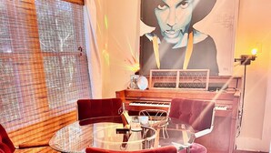 Uma sala de jantar com mesa de vidro rodeada por cadeiras estofadas em vermelho, um piano e um grande quadro abstrato na parede.