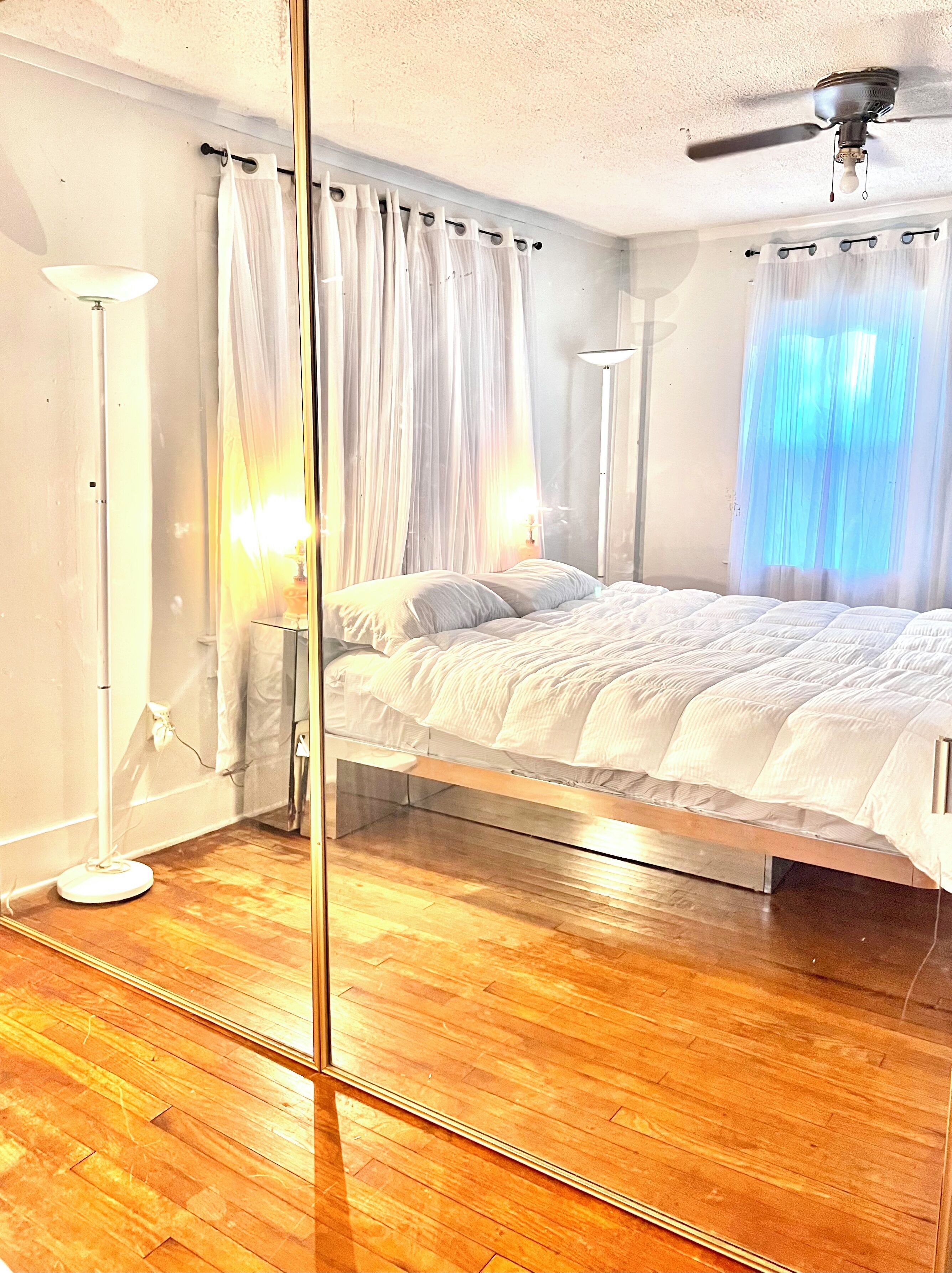 A bedroom with a bed, a standing lamp, a ceiling fan, and sheer curtains.