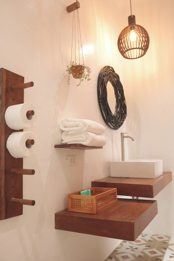 Een badkamer met een houten handdoekhouder, toiletpapier, een plank met opgevouwen handdoeken, een hanglamp en een wastafel.