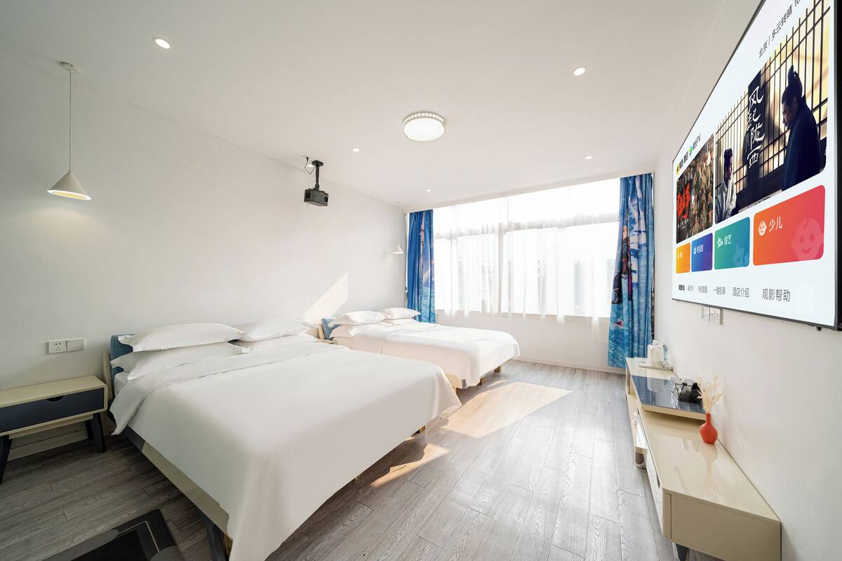 A hotel room with two beds, a desk, a projector, and a large window with curtains.