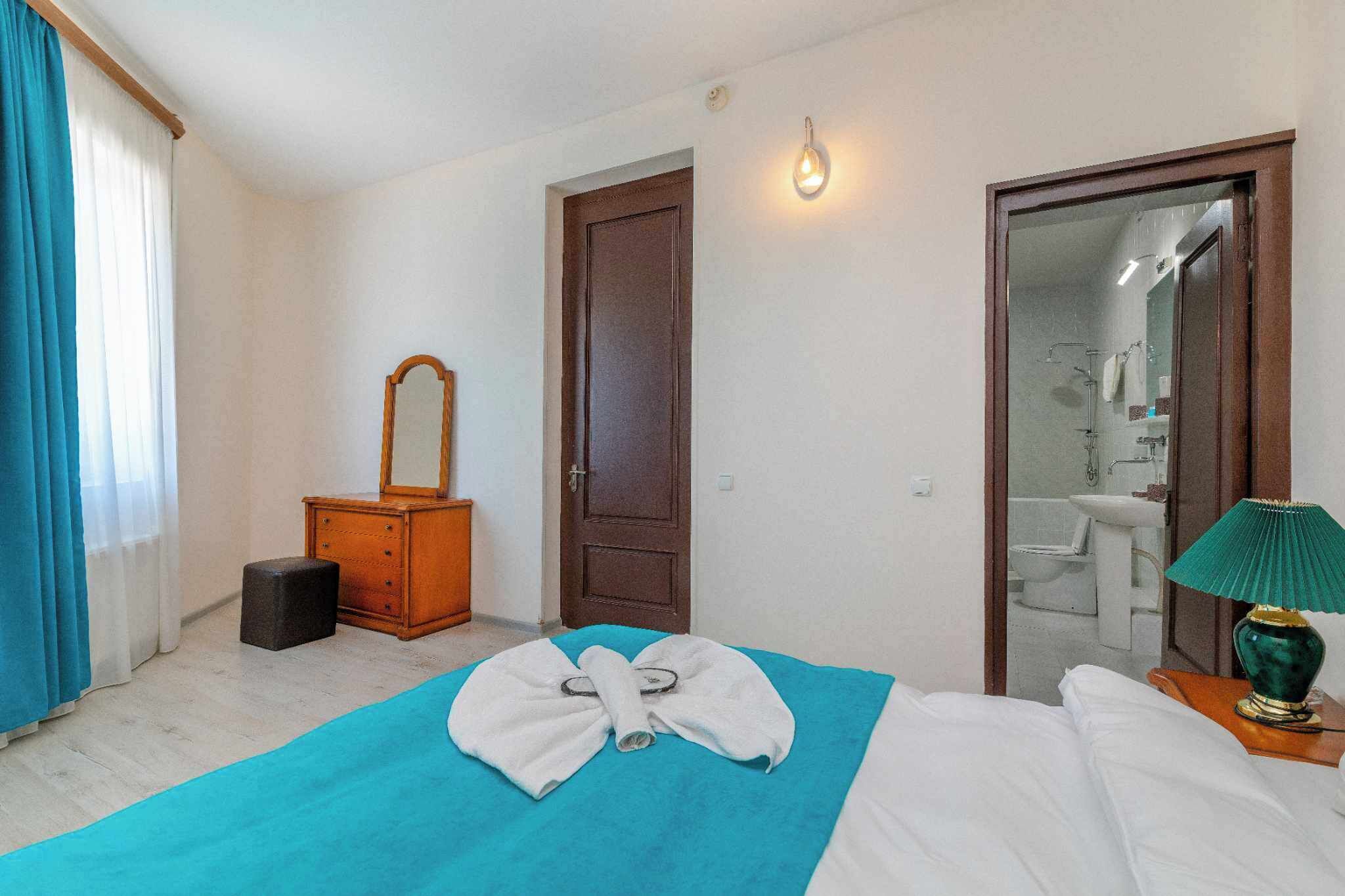 Hotellihuone, jossa on sänky, puinen lipasto, peili, avoimesta ovesta näkyvä kylpyhuone ja vihreä lamppu yöpöydällä.