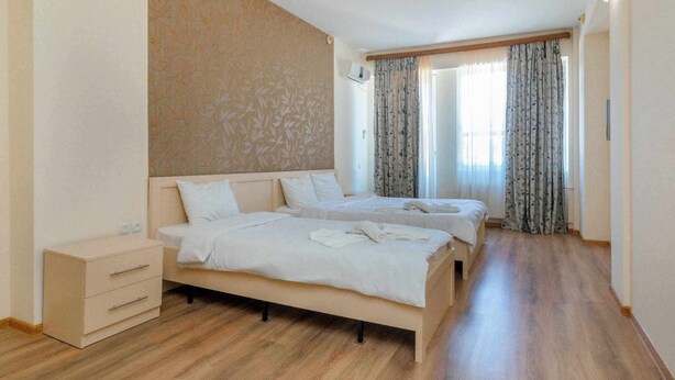 A hotel room with two beds, a wooden floor, a nightstand, and a window with curtains. - Victoria Hotel (Gori)