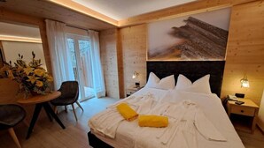 A bedroom with a large bed, a wooden headboard, and a painting above it. There is a small table with a chair and a vase of flowers. - Lake & Nature Hotel Gloria (Molveno)