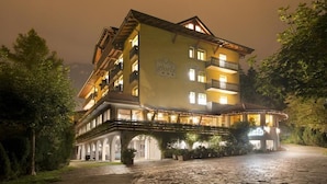 A multi-story hotel with a balcony on the second floor, a covered outdoor area with plants, and a stone-paved entrance. - Lake & Nature Hotel Gloria (Molveno)