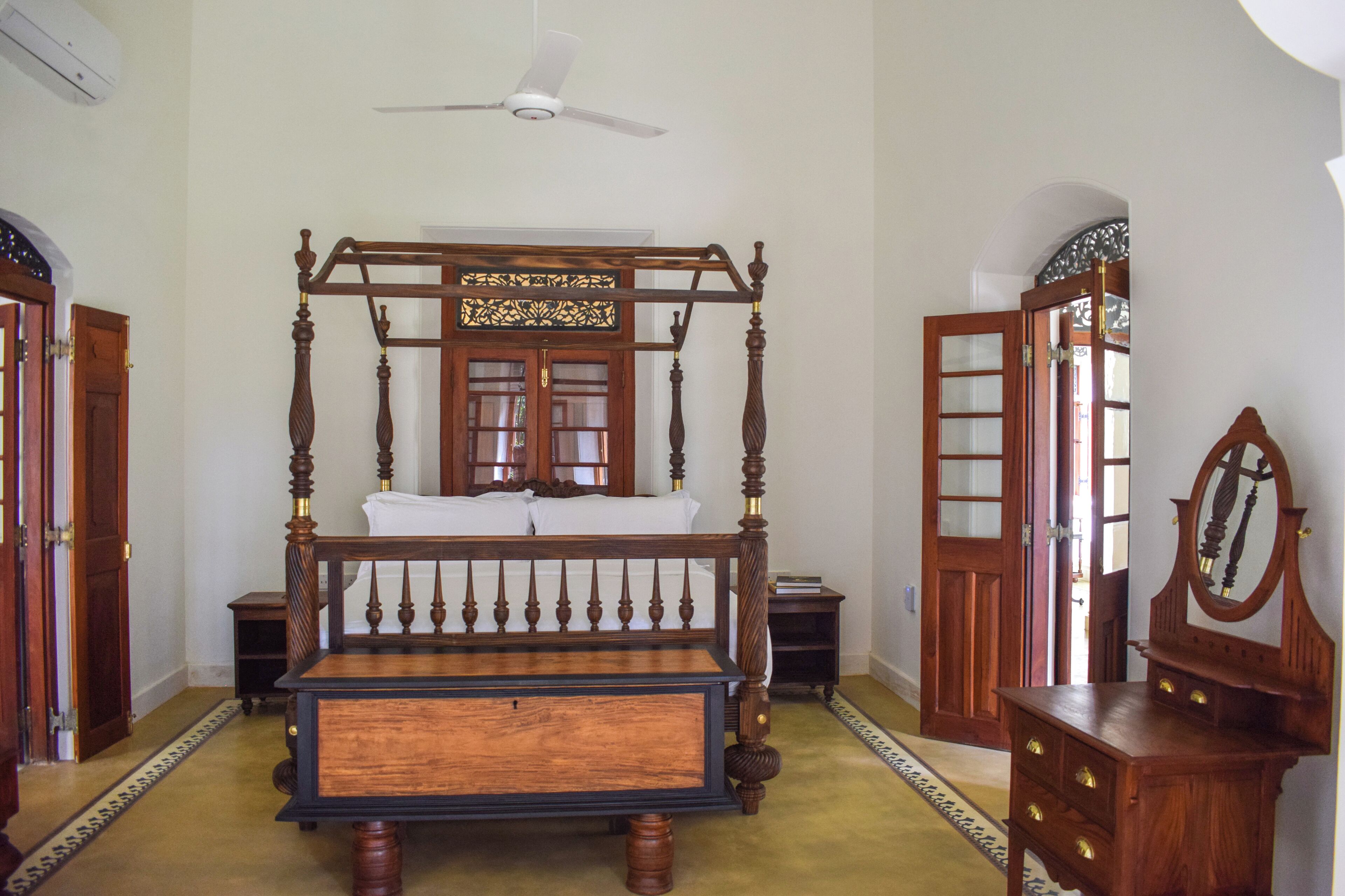 A four-poster bed with a wooden frame and a headboard with a decorative design. There is a ceiling fan above the bed, and a wooden dresser with a mirror to the right.