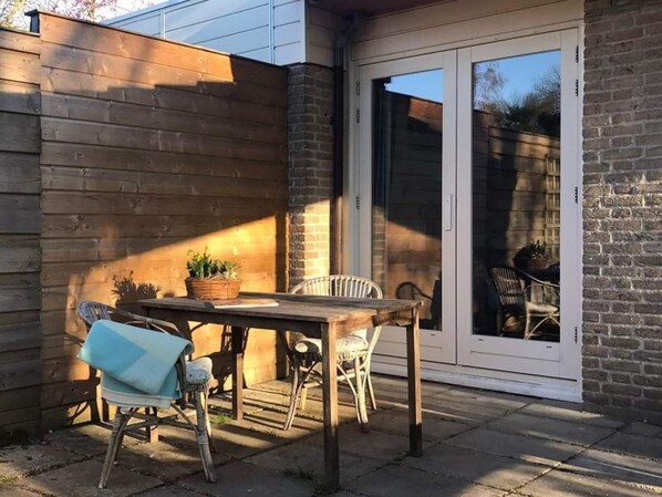 Une terrasse avec une table et des chaises en bois, un panier de plantes et une porte-fenêtre coulissante.