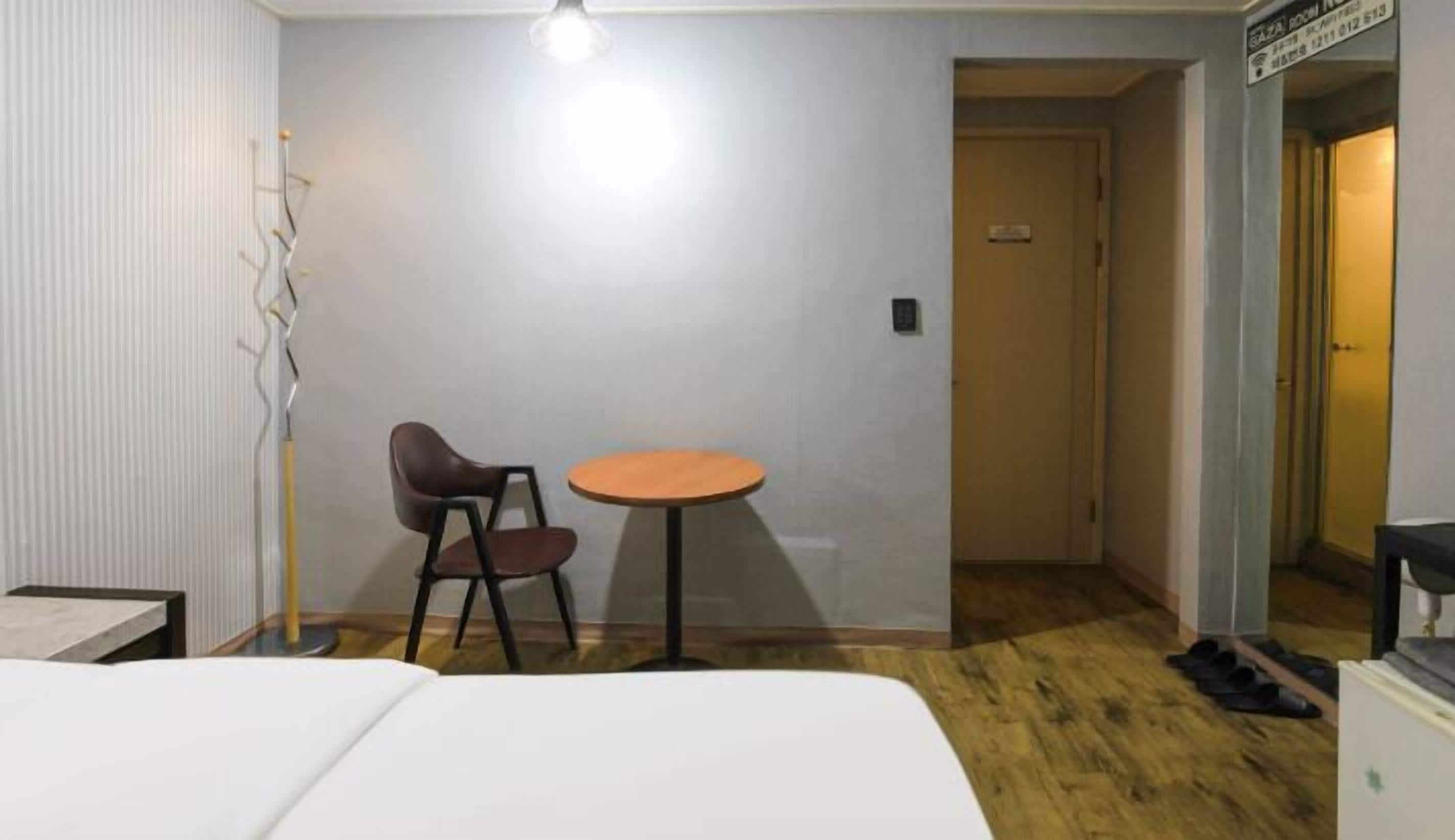 A small, simple room with a bed, a chair, a small table, and a door leading to another room.