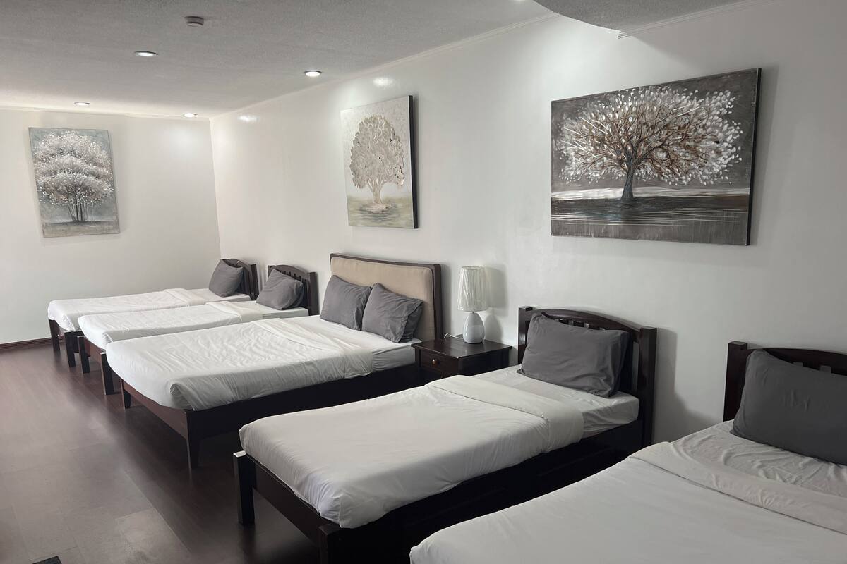 A hotel room with three single beds, a nightstand with a lamp, and artwork on the walls.