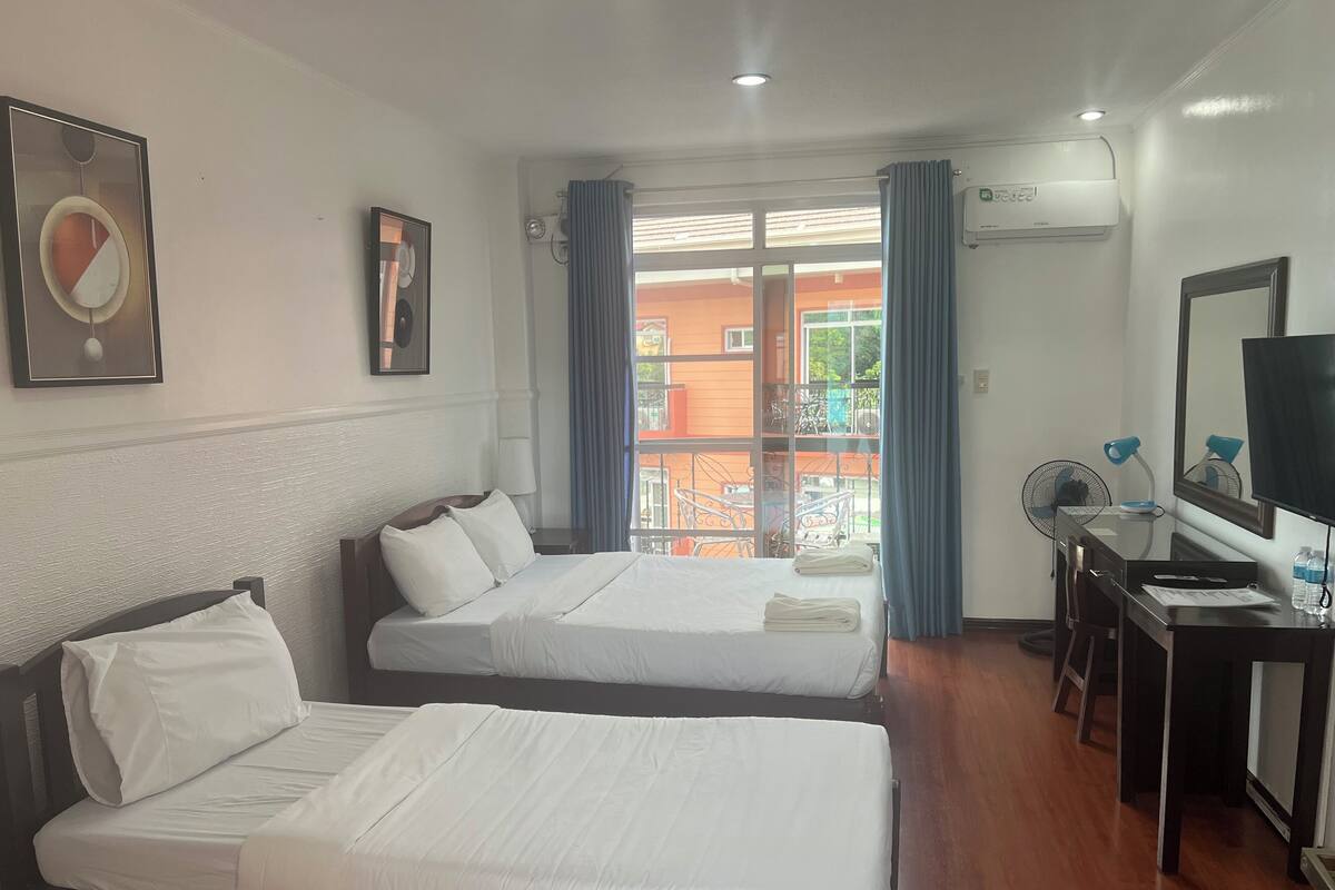 A hotel room with two beds, a desk with a computer, a TV, and a balcony with outdoor furniture.