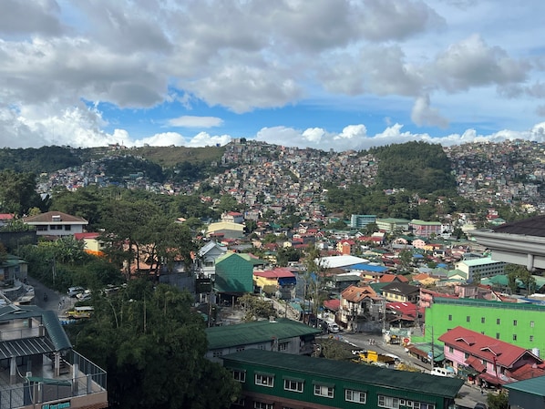 A densely packed hillside town with a mix of residential and commercial buildings, a prominent green building in the foreground, and a clear sky with scattered clouds. - NYC Manhattan Suites (Baguio)