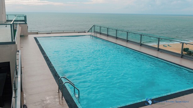 Una piscina rettangolare con ringhiera in vetro, adiacente a una spiaggia con spiaggia sabbiosa e vista sull'oceano.