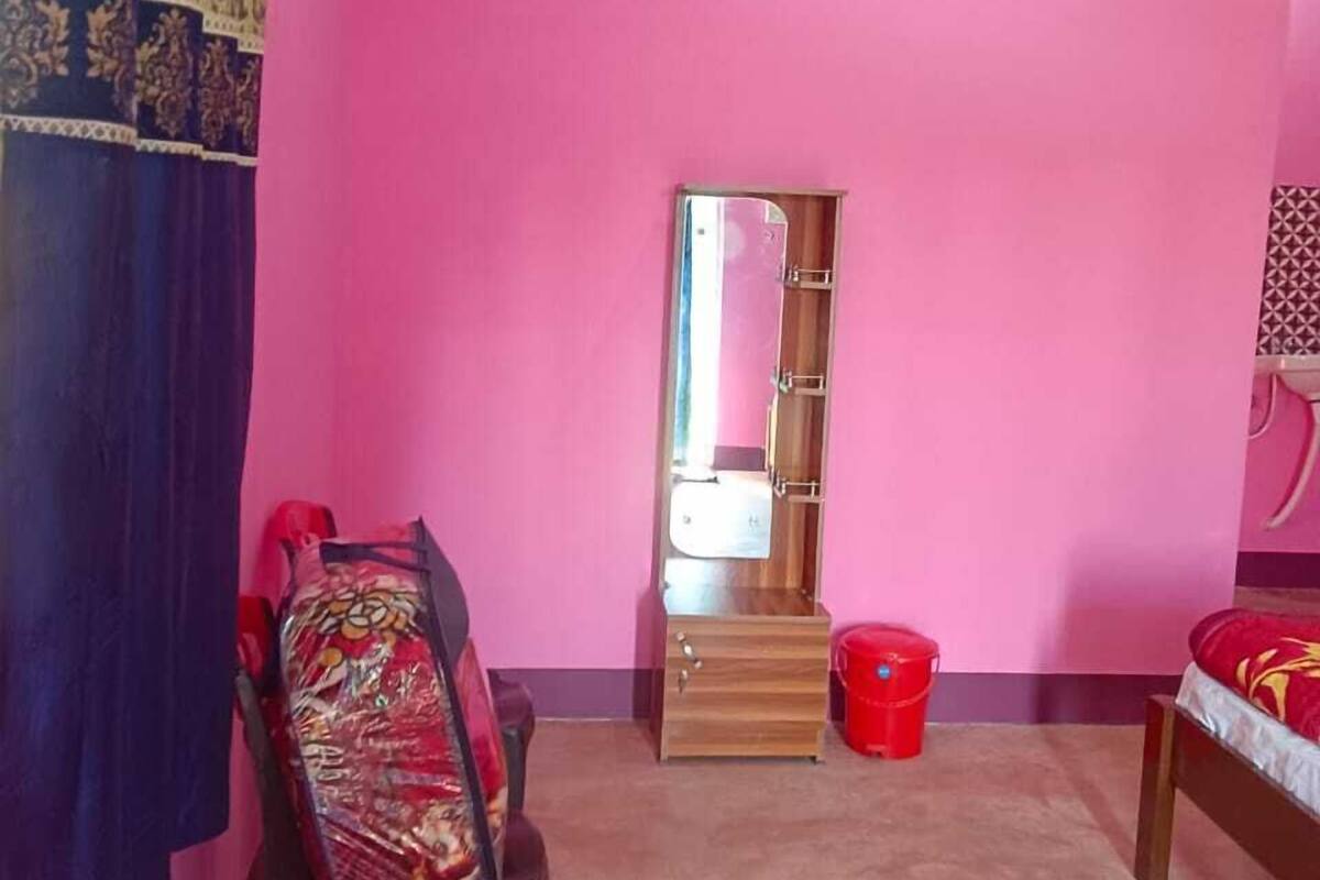 A pink-walled room with a wooden wardrobe, a chair with a patterned cushion, a red bucket, and a bed with a red and white blanket.