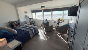 A bedroom with a bed, a chair, a table with breakfast items, a TV, and a view of the sea.