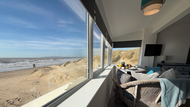 A room with a large window overlooking a beach, a wicker sofa, a flat-screen TV, and a dining table with breakfast items.