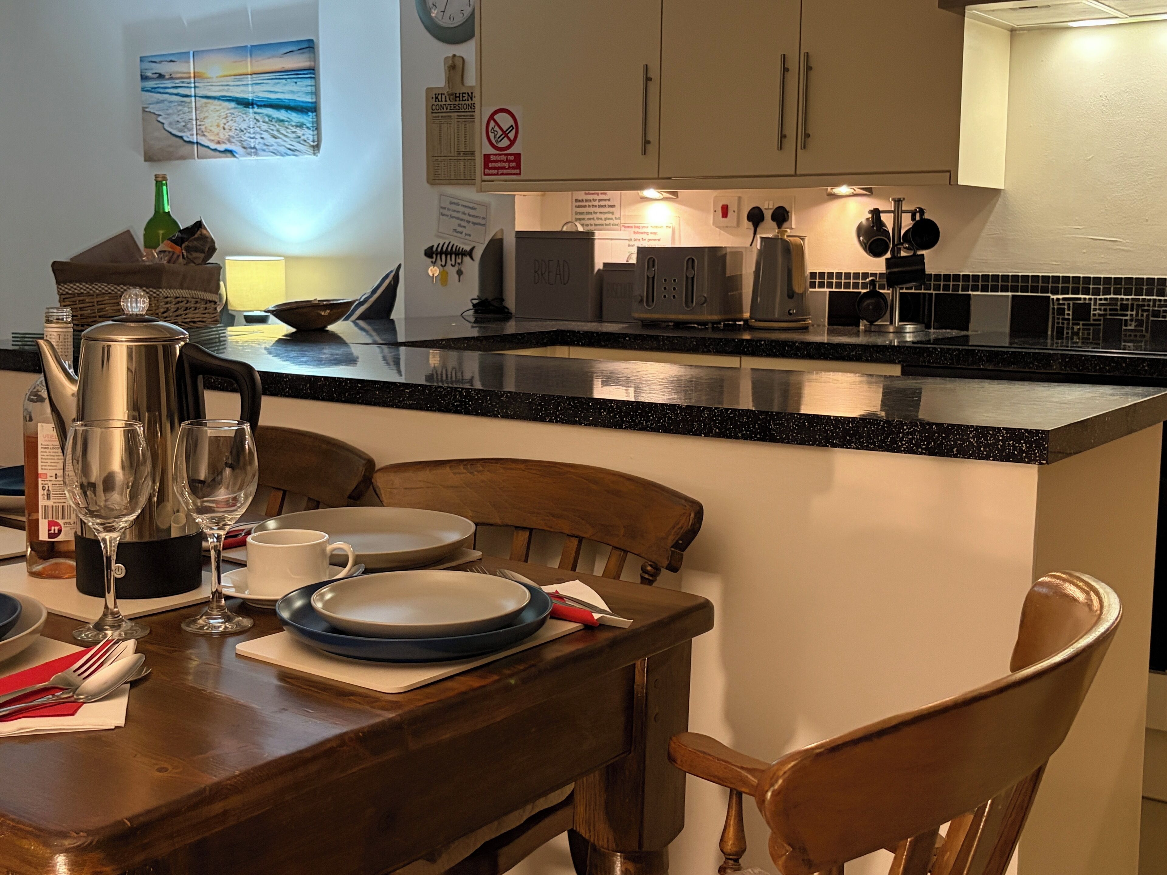 A kitchen with a dining area, wooden chairs, a table set with plates, glasses, and cutlery.