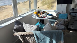 A corner room with a large window overlooking a beach, a dining table set with breakfast, and a TV on the wall.