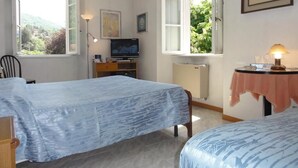 A room with two beds, a television, a desk with a lamp, and a window with a view of greenery. - Albergo Ristorante Grigna (Mandello del Lario)