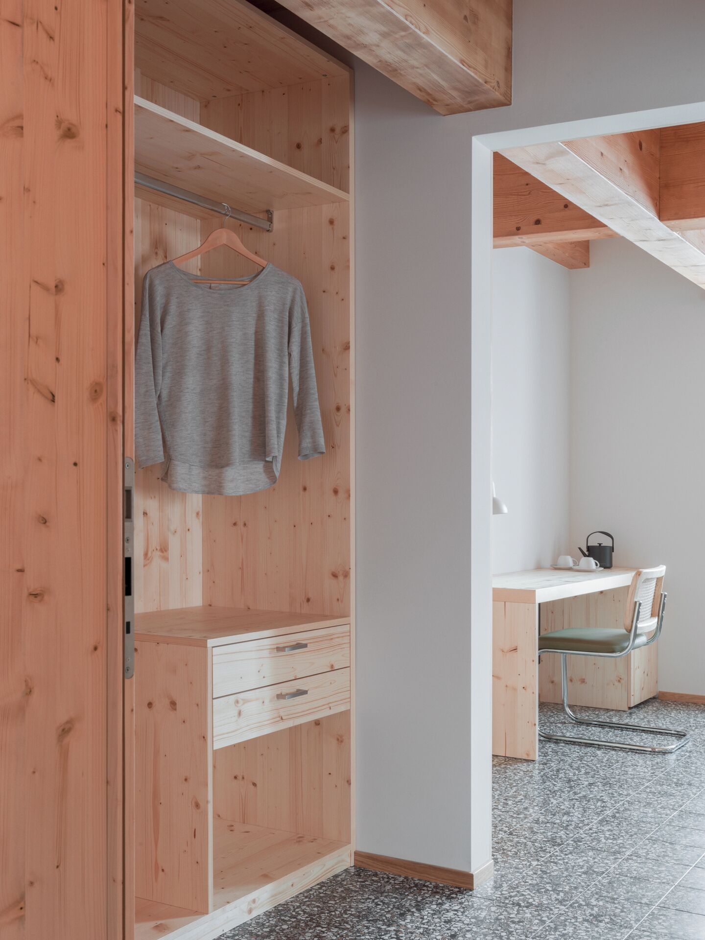A wooden wardrobe with a hanging shelf, a desk with a chair, and a tiled floor.