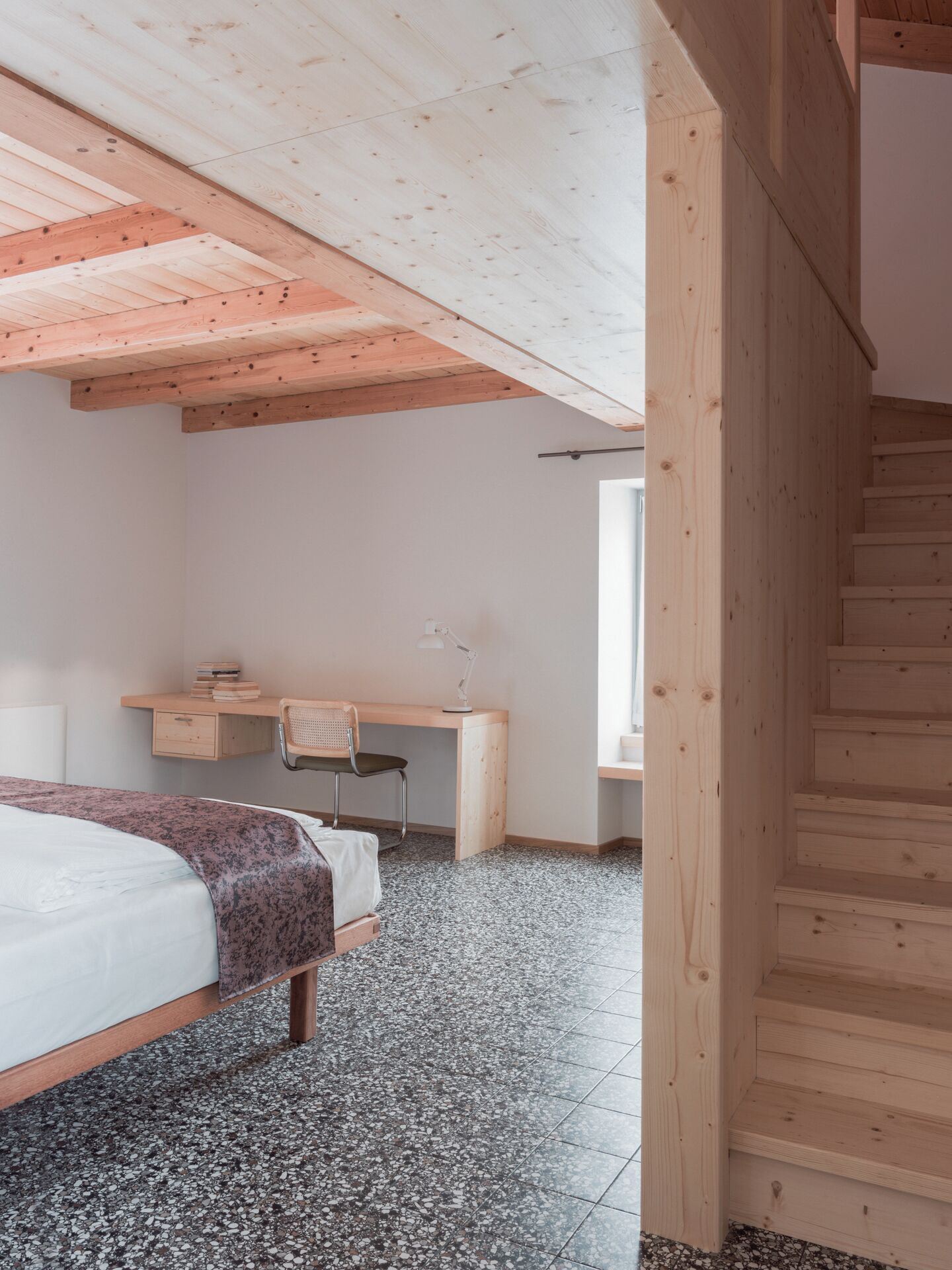 A bedroom with a bed, a desk, and a chair. There is a staircase leading to an upper level.