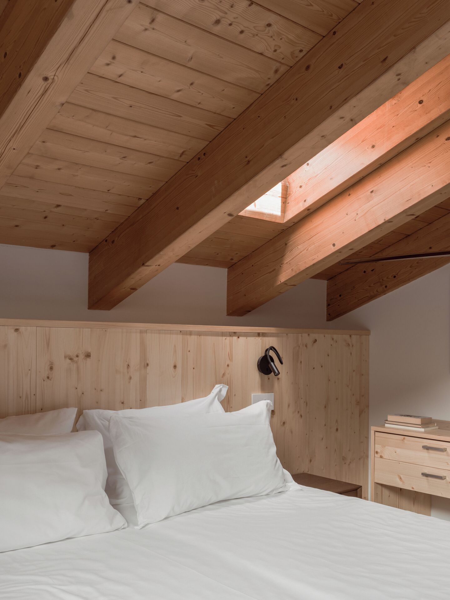 A bedroom with a wooden ceiling, a bed with white bedding, a bedside table, and a wall-mounted lamp.