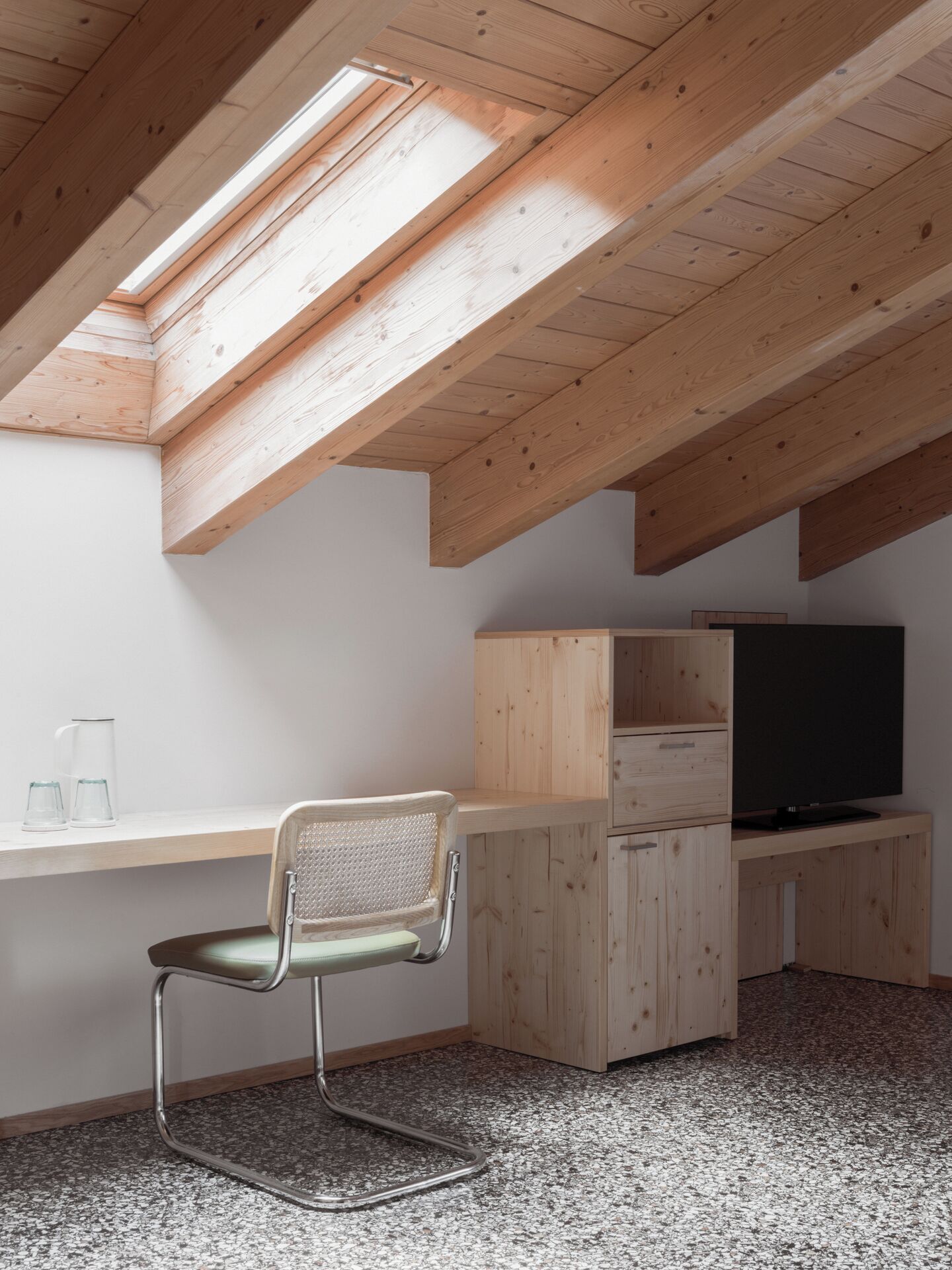Una stanza con soffitto spiovente, travi in legno, una scrivania con una sedia, un televisore e un dispenser d'acqua.
