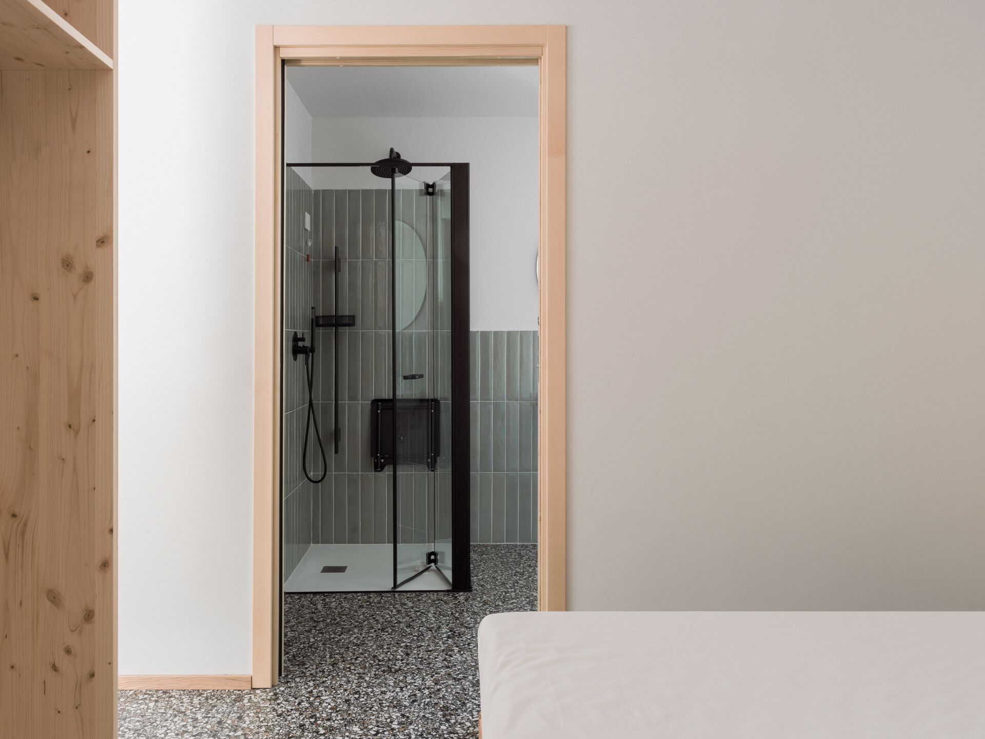 Ein Zimmer mit Holztür, das in einen Duschbereich mit Glasabtrennung führt, mit einer kleinen schwarzen Steuerung und einem gefliesten Boden.