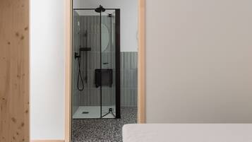 Ein Zimmer mit Holztür, das in einen Duschbereich mit Glasabtrennung führt, mit einer kleinen schwarzen Steuerung und einem gefliesten Boden.