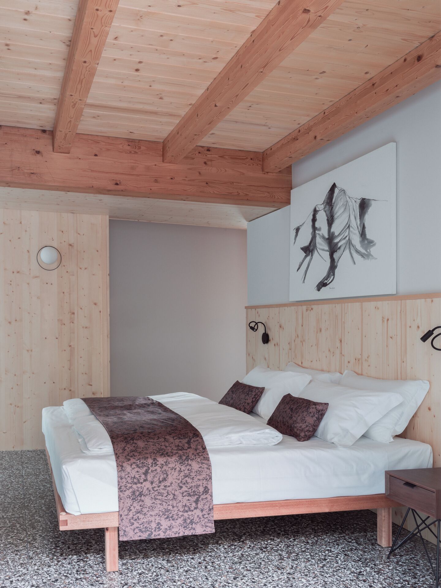 A bedroom with a wooden ceiling, a bed with white and brown bedding, a headboard with a light fixture, and a wall-mounted artwork.