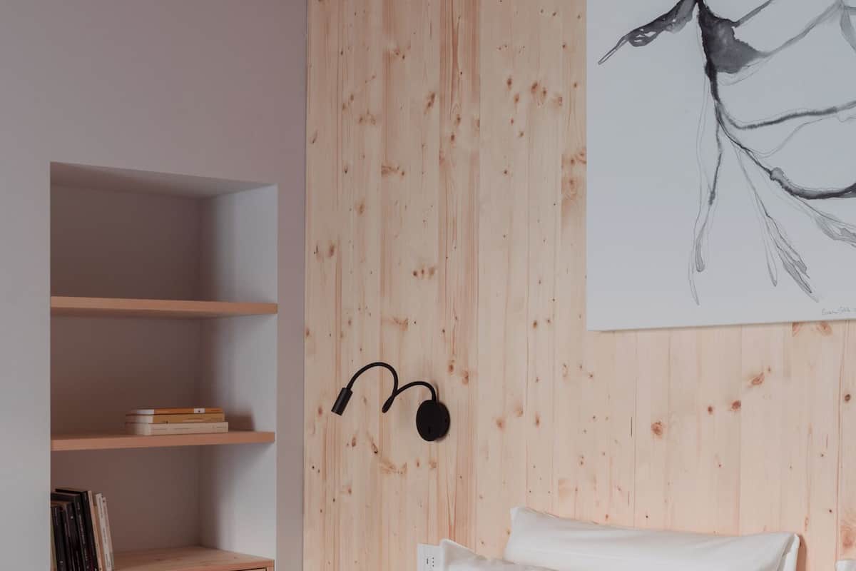 Ein Schlafzimmer mit Holzbalkendecke, einem Bett mit weißer Bettwäsche, einem Wandregal mit Büchern und einem großen abstrakten Gemälde.