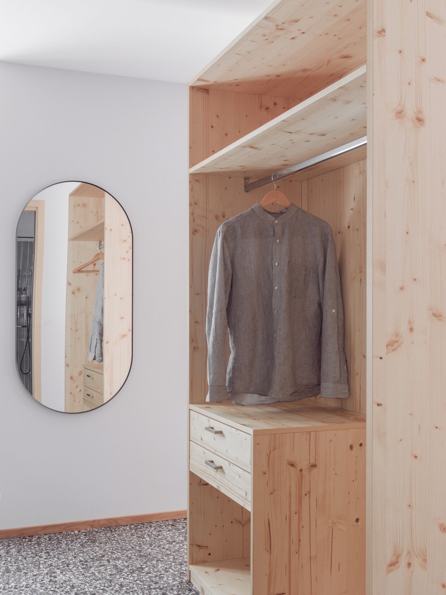 Ein hölzerner Kleiderschrank mit Kleiderstange und Schubladen, ein Spiegel an der Wand und ein grau gestreiftes Hemd darin hängend.