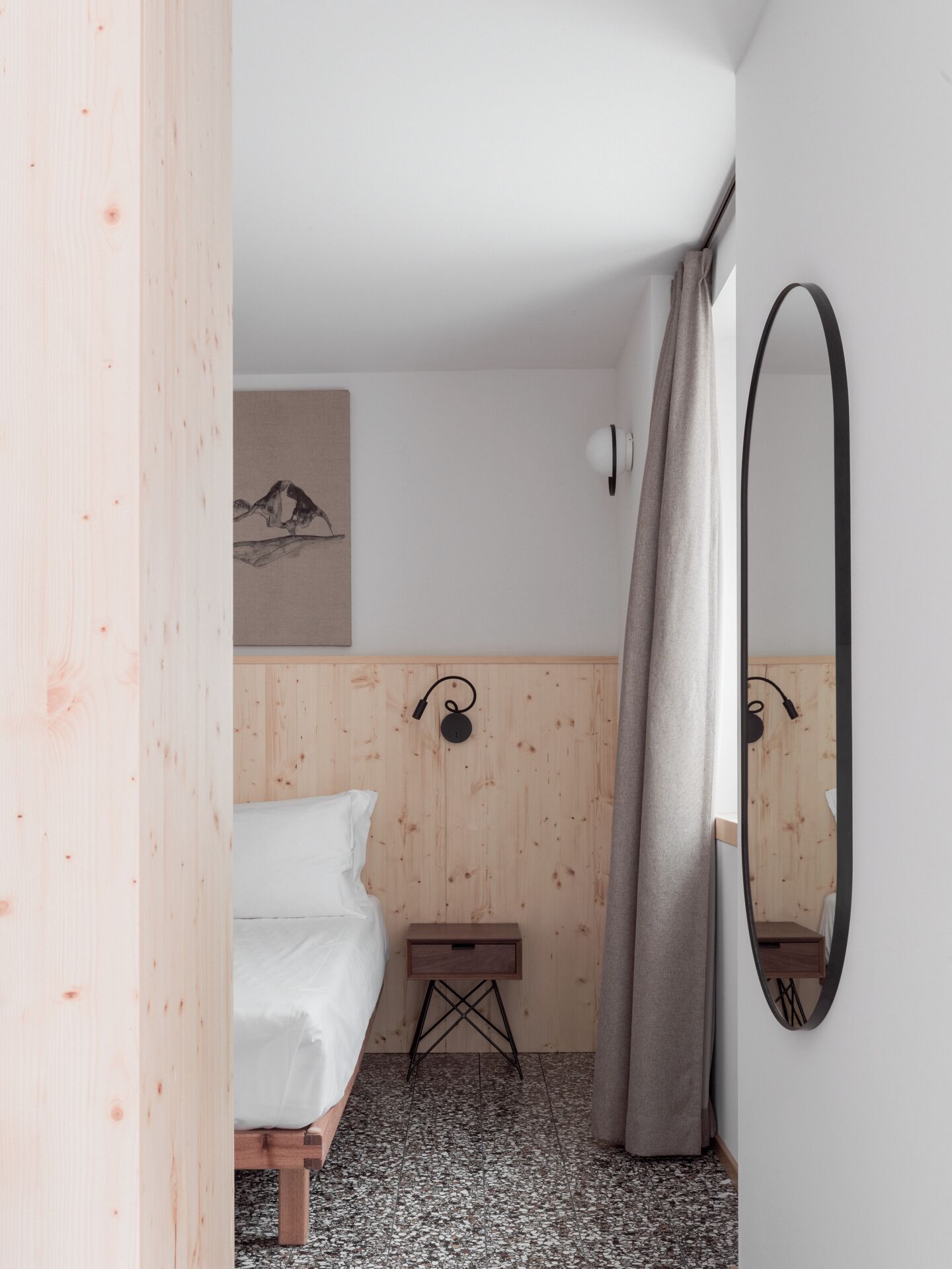 A bedroom with a bed, a small bedside table, a wall-mounted lamp, a mirror, and a wooden wall panel.
