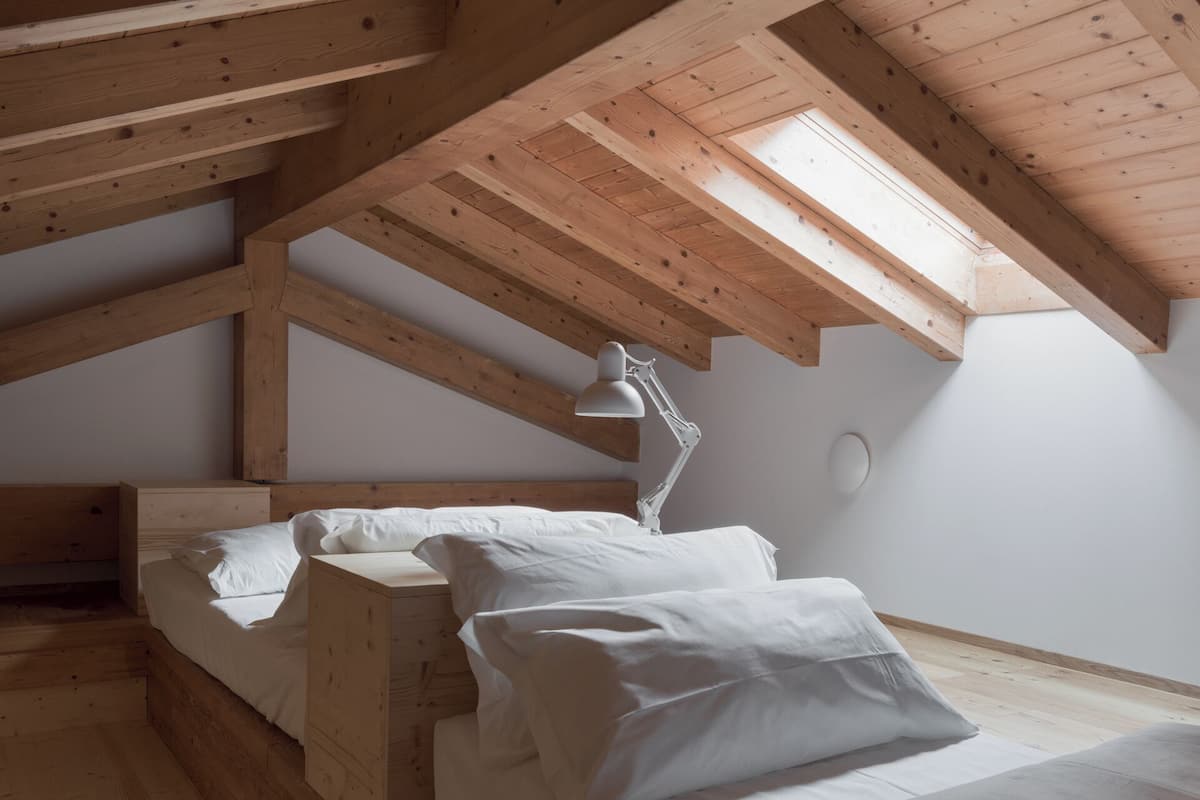 Ein Schlafzimmer mit geneigter Decke, Holzbalken, einem Oberlicht, einem Bett mit weißer Bettwäsche und einem Schreibtisch mit Lampe.