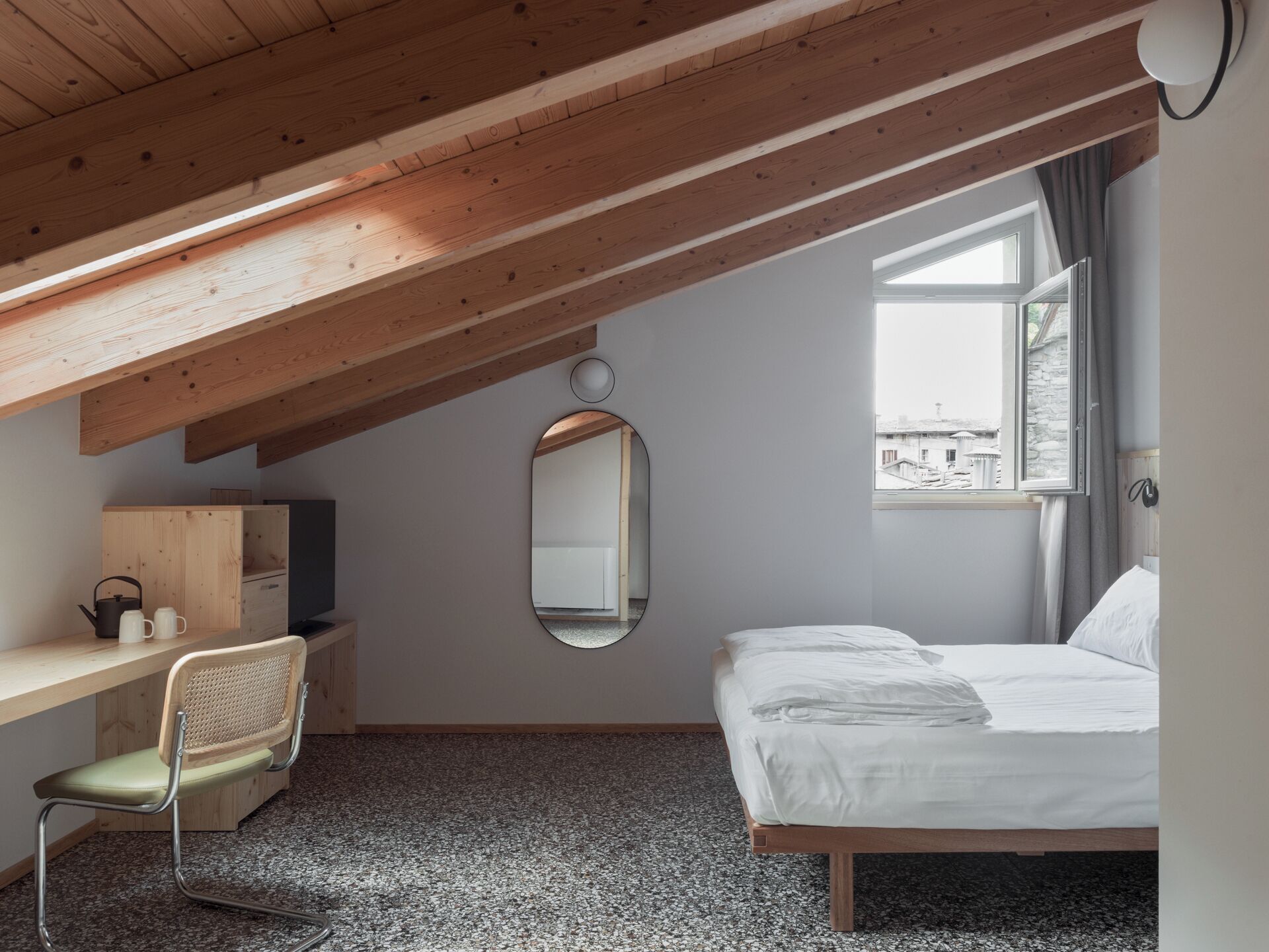 A bedroom with a sloped ceiling, a bed, a desk with a chair, a mirror, and a window with a view of buildings.