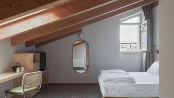 A bedroom with a sloped ceiling, a bed, a desk with a chair, a mirror, and a window with a view of buildings.