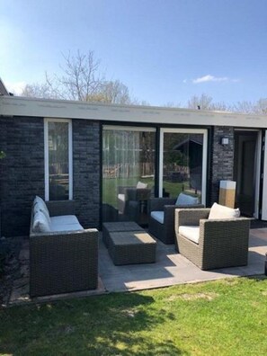 A modern outdoor patio with wicker furniture and glass doors leading to a backyard. - Furnished Bungalow Near Beach (Sint Maartensvlotbrug)