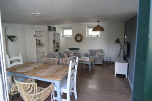 A spacious living room with a wooden dining table, white chairs, a grey sofa, and a white cabinet. - Furnished Bungalow Near Beach (Sint Maartensvlotbrug)