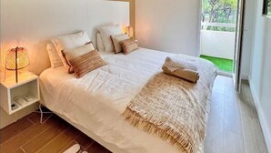 A modern bedroom with a large bed, a bedside table with a lamp, and a view of the outdoors through a sliding glass door. - Saint-Raphaël - Apartment near Valescure golf course and sea.  (Saint-Raphaël)