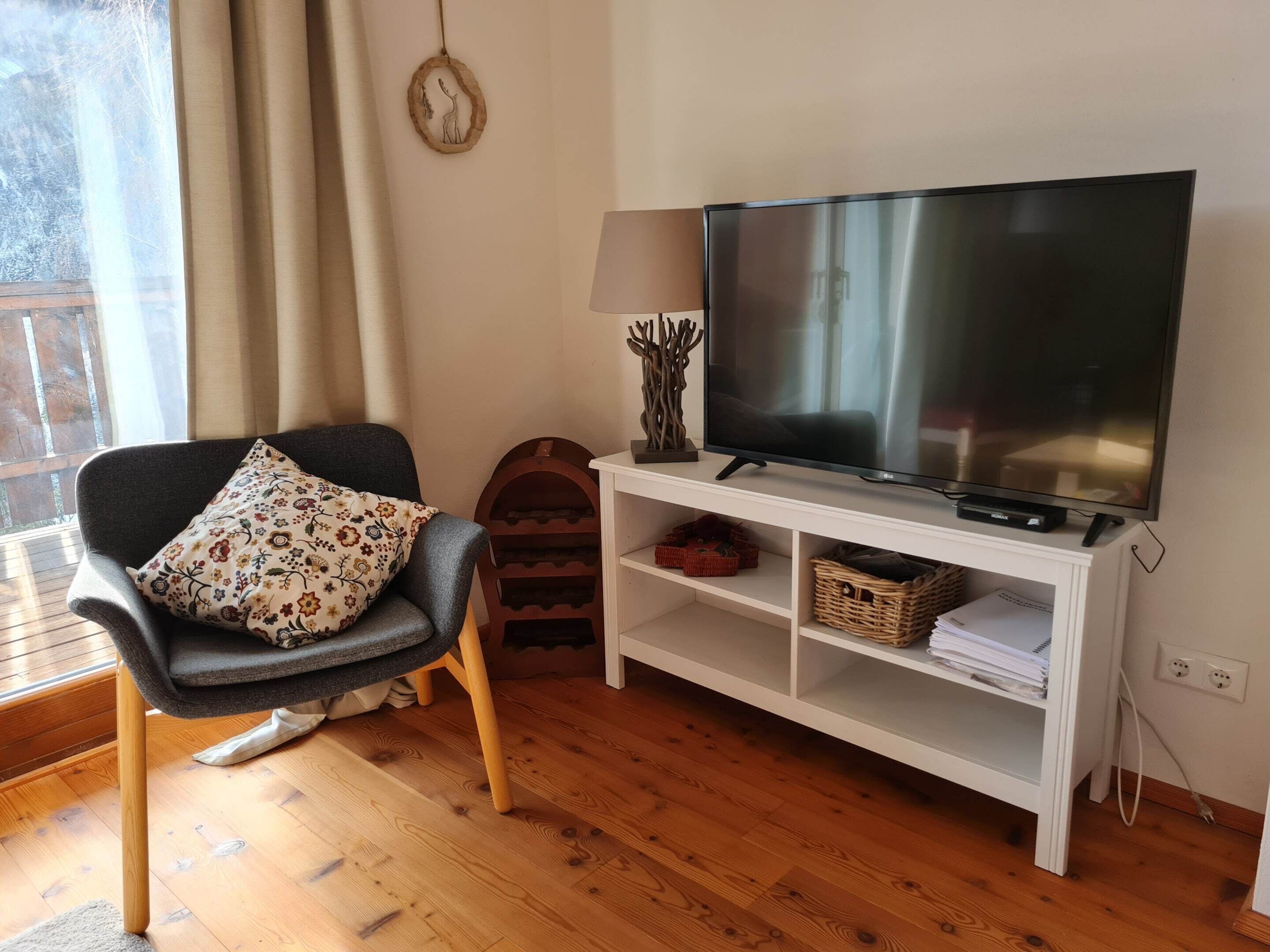 Un salón con un sillón gris, un televisor de pantalla plana sobre un soporte blanco, piso de madera y una ventana con cortinas.