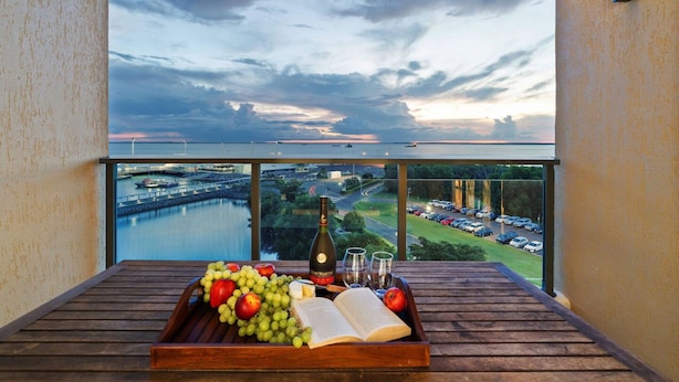 Ein Balkon mit Holztisch, gedeckt mit Trauben, Tomaten, einer Weinflasche und Gläsern, mit Blick auf ein Gewässer und eine Brücke.