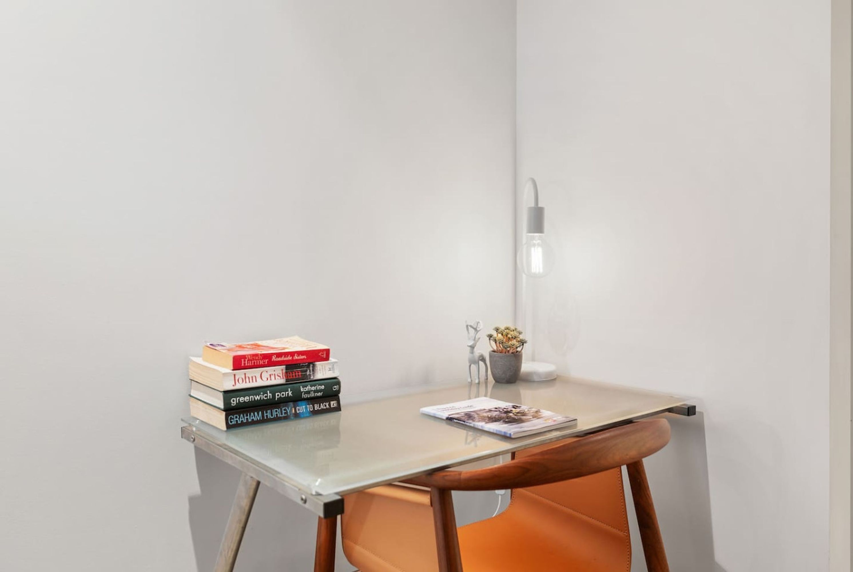 A small, modern workspace with a glass table, a stack of books, a decorative plant, and a wall-mounted light.