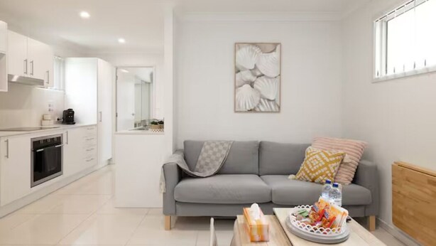 A modern living room with a grey sofa, a coffee table with snacks, and a kitchen area with white cabinets and appliances. - Smart Suites Self Check-in Apartments - 35 Freney (Rocklea)