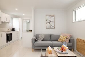 Een moderne woonkamer met een grijze bank, een bijzettafeltje met lekkernijen, en een keuken met witte kasten en apparatuur.