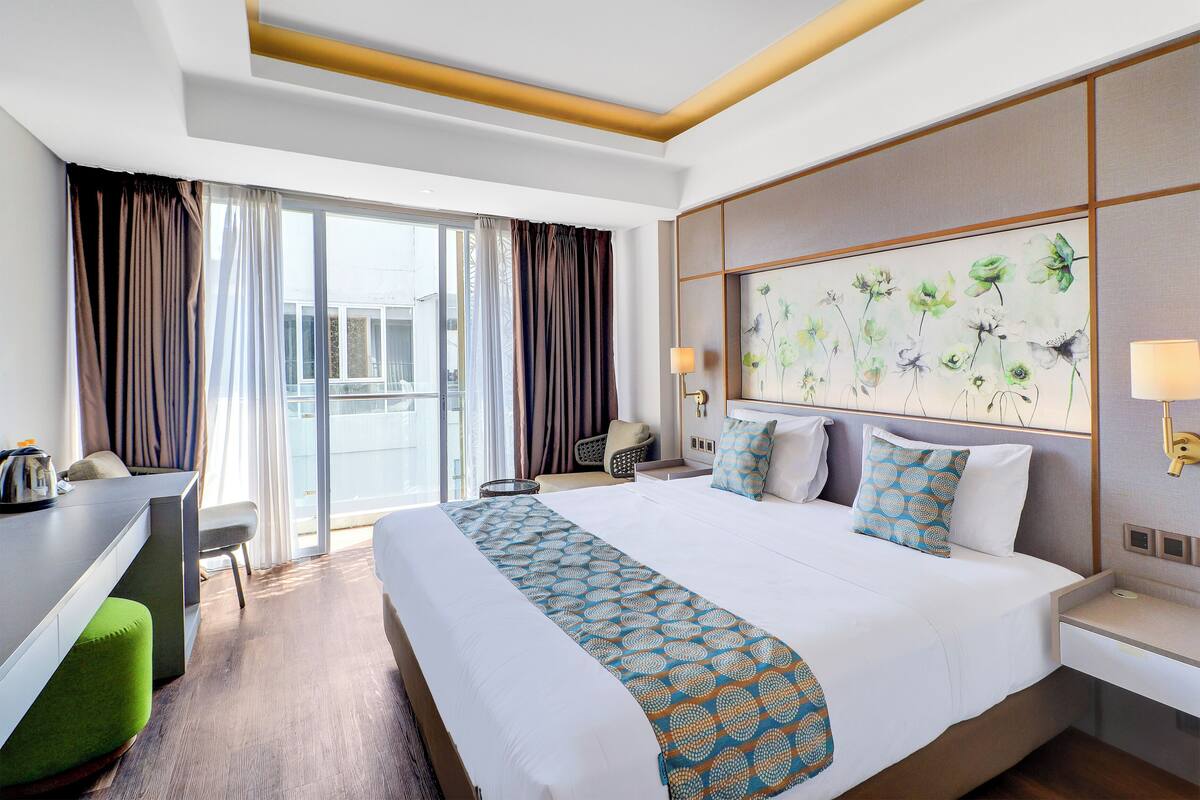 Une chambre d’hôtel avec un grand lit, un bureau et une chaise. Il y a une fenêtre avec des rideaux, un tableau au mur et une lampe sur la table de chevet.