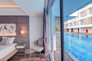 A modern hotel room with a large window, a sofa, a small table, and a view of the pool. - Sunday Hotel Arshika Sunset Road (Kuta)