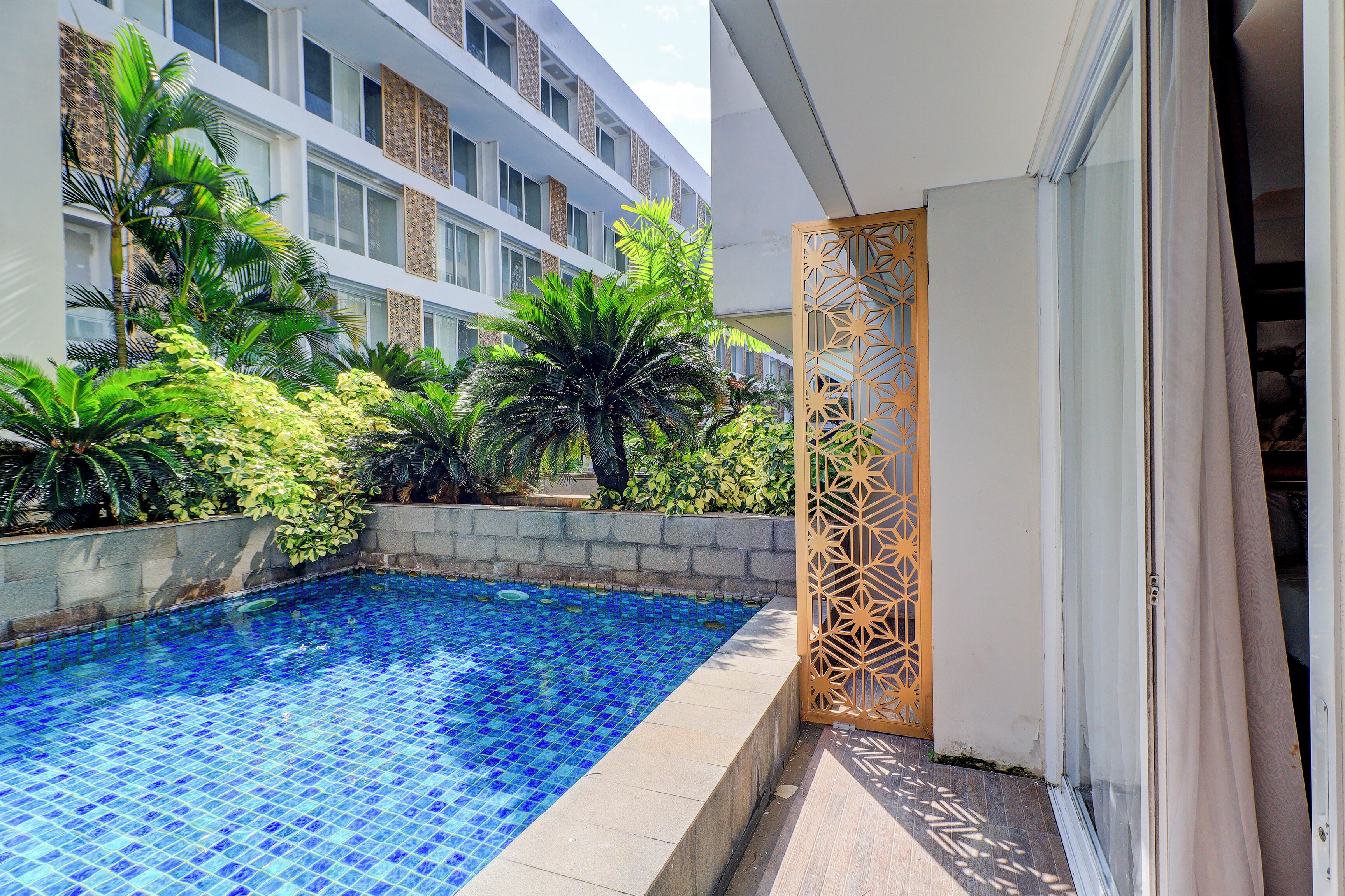 Una piscina con azulejos azules, rodeada de vegetación y un edificio con múltiples ventanales.