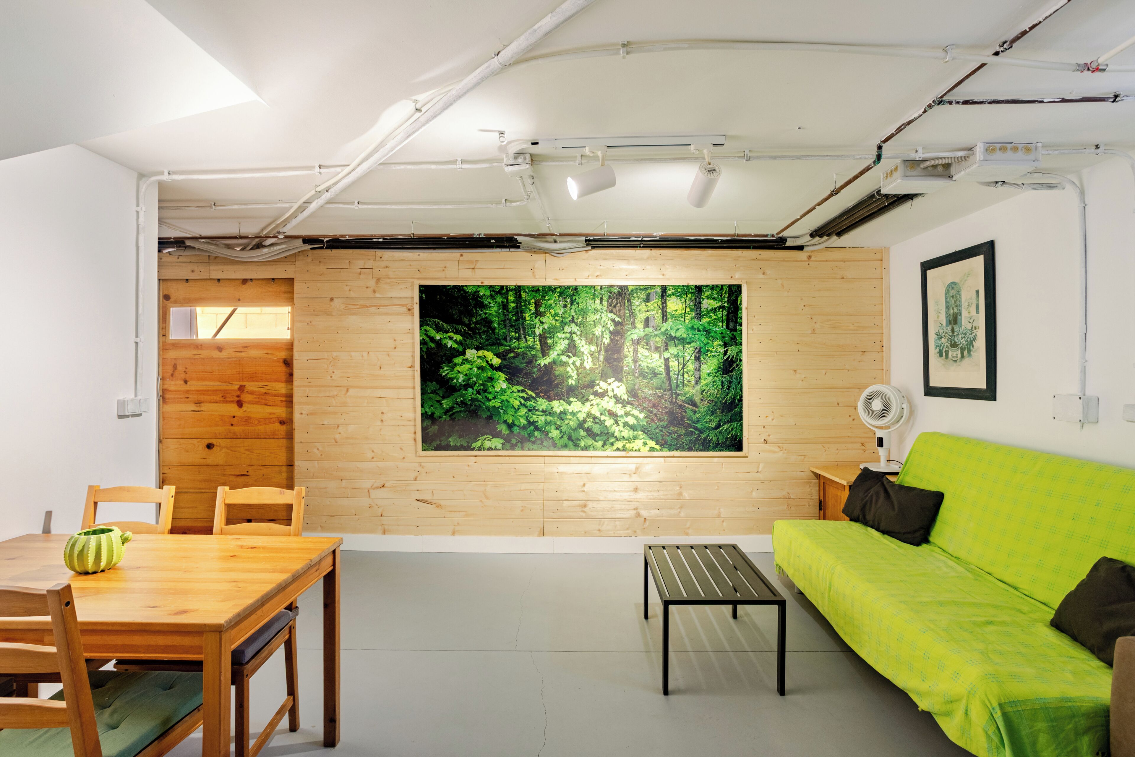 Une pièce avec un mur en bois, une grande fresque représentant une forêt, un canapé vert, une table en bois avec des chaises et un petit ventilateur.