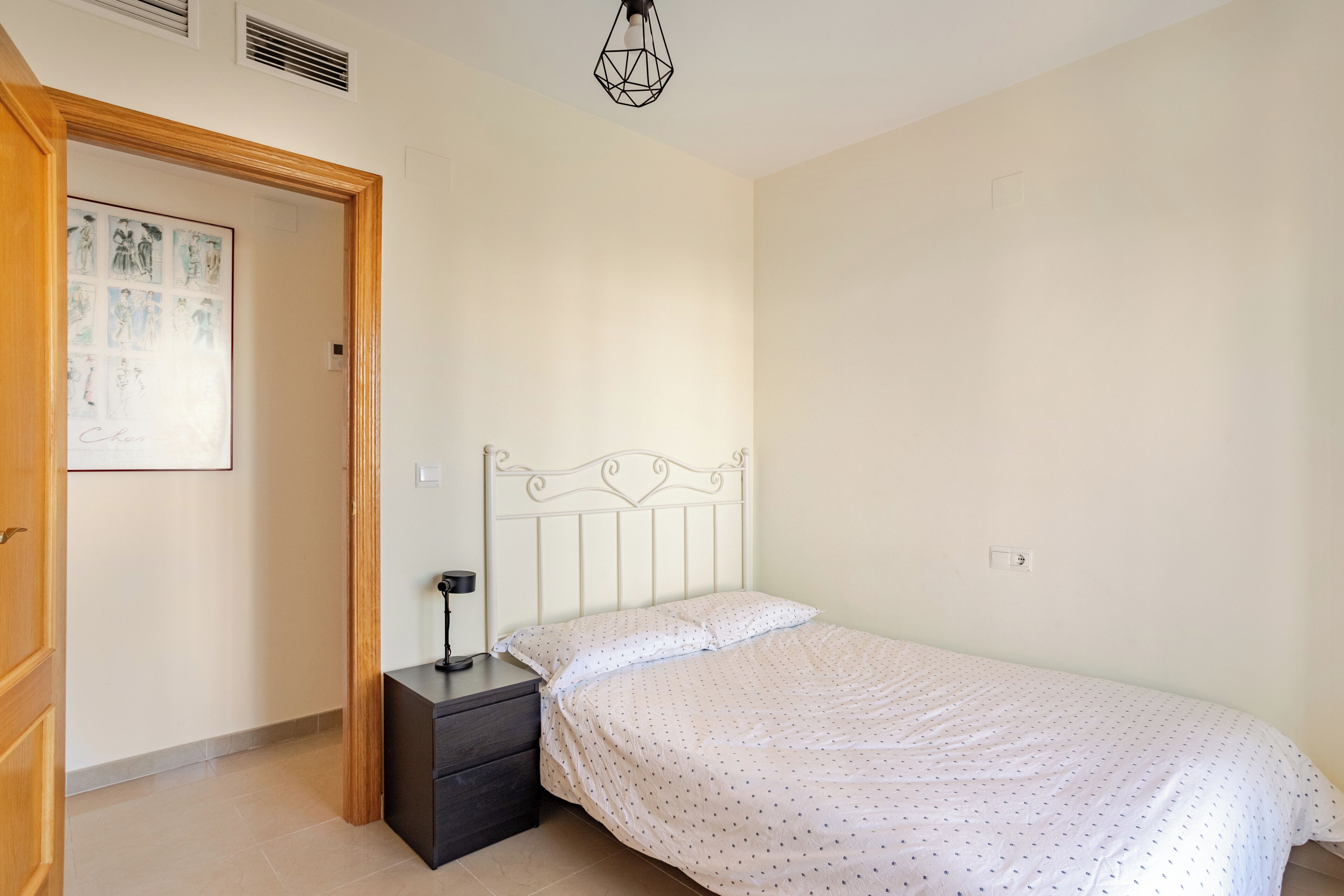 A bedroom with a bed, a nightstand, a wall-mounted lamp, and a door leading to another room.