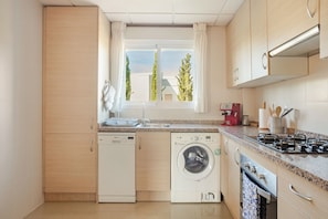 Une cuisine compacte équipée d’un lave-linge, d’un lave-vaisselle, d’une cuisinière et d’une fenêtre donnant sur les arbres.