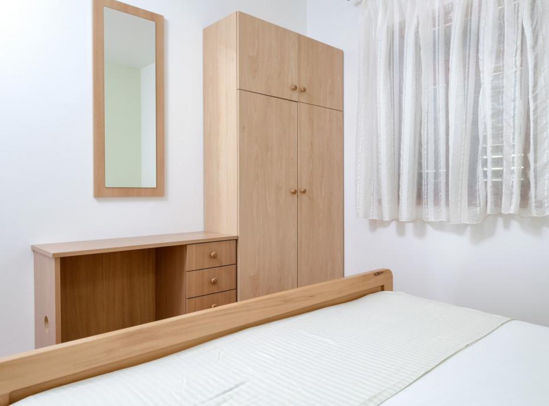A bedroom with a bed, a wooden wardrobe, a mirror, and a window with sheer curtains.